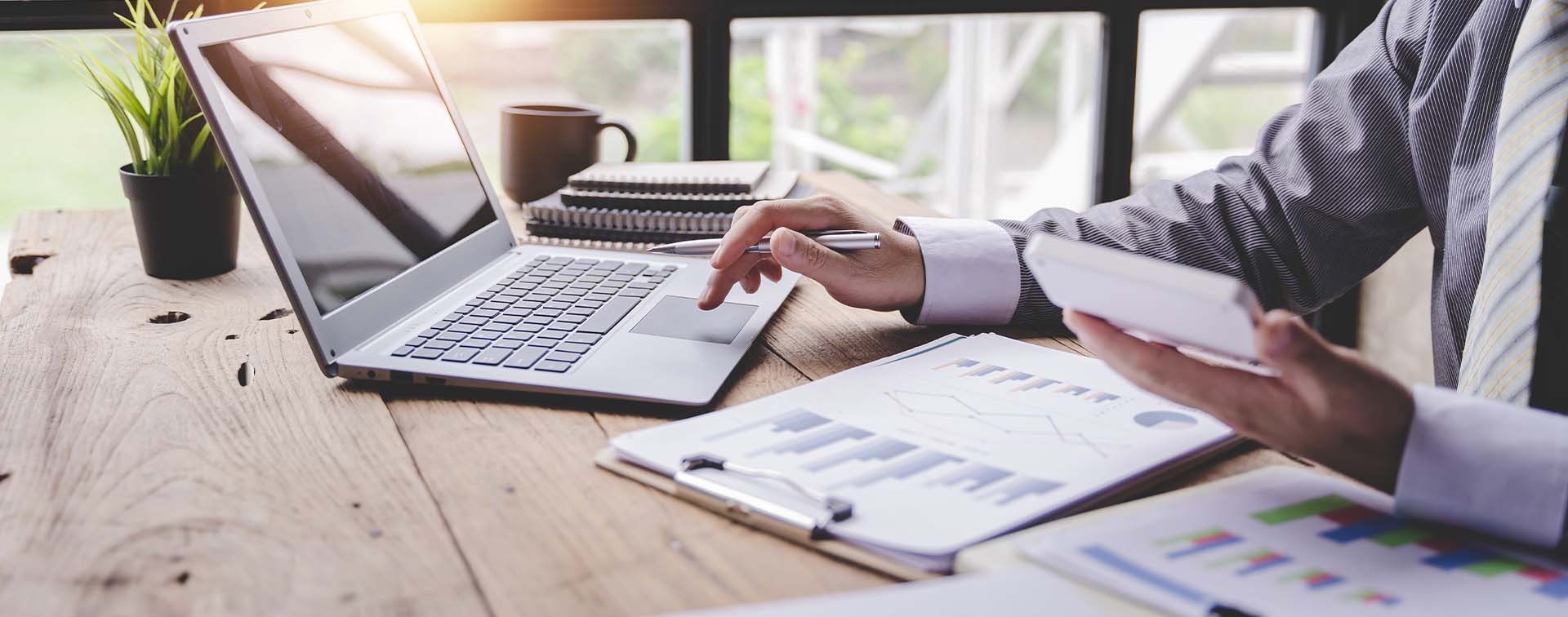 Closeup image of a person analyzing charts on a desk and scrolling on a laptop with a calculator in one hand