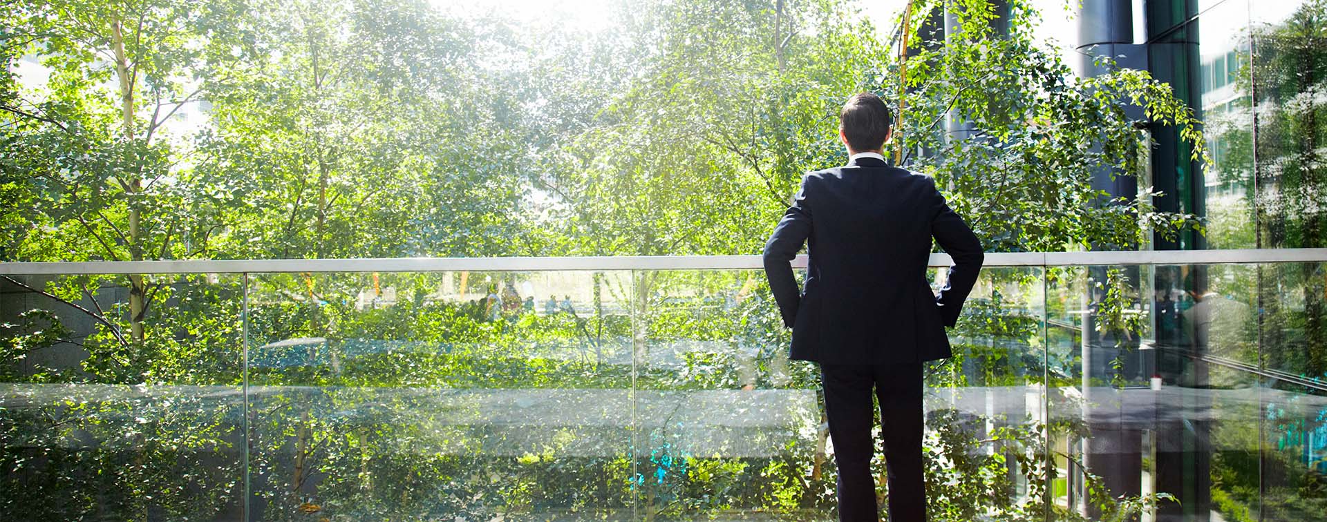 Businessman on a glass balcony looking at trees contemplatively